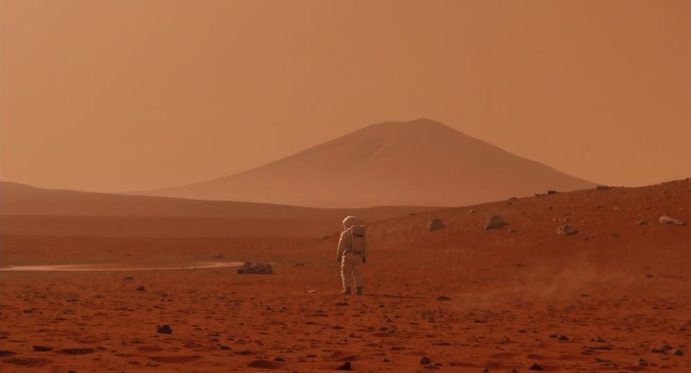 An image of a lone astronaut standing on the surface of Mars, looking at a distant mountain.png